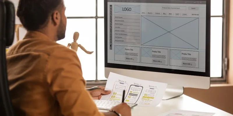 Profesional diseñando un sitio web con prototipos en papel y maqueta digital en monitor.
