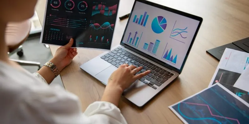 Persona analizando gráficos financieros en laptop y hojas impresas sobre escritorio.